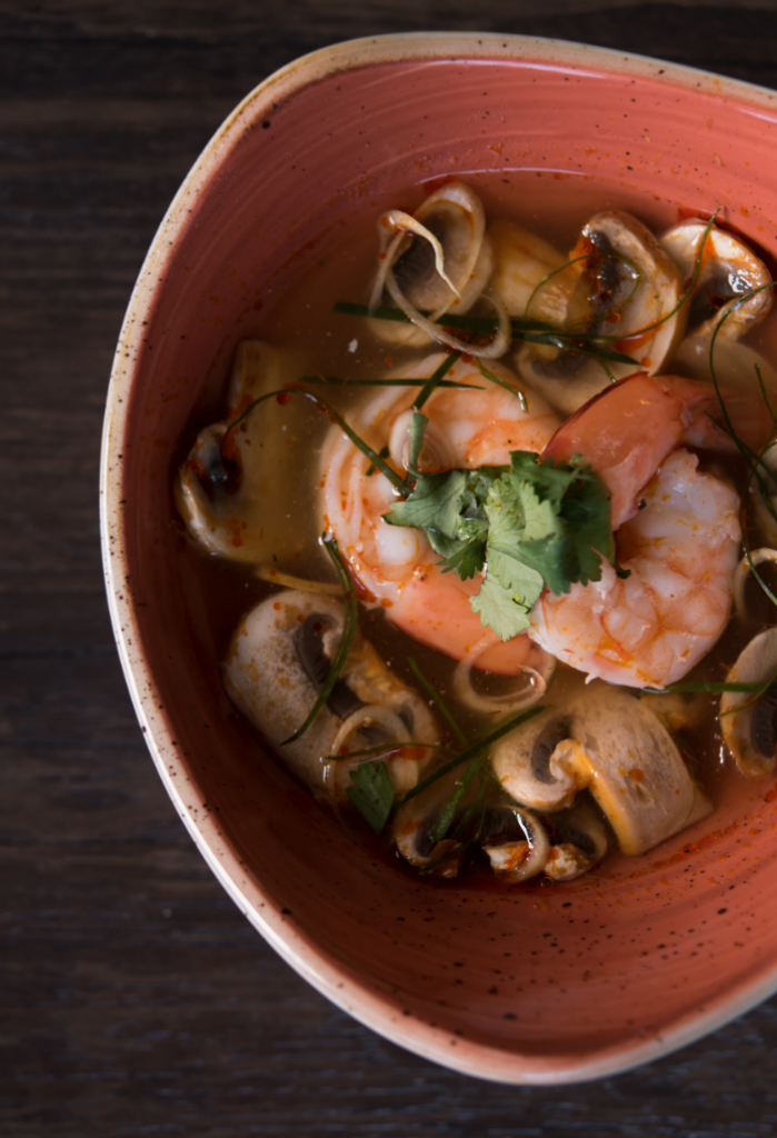 Overhead of a triangular red bowl with Tom Yum Soup prepared with with lemon grass, galangal, kaffir, mushroom, lime leaves and lime juice topped with cooked shrimp.