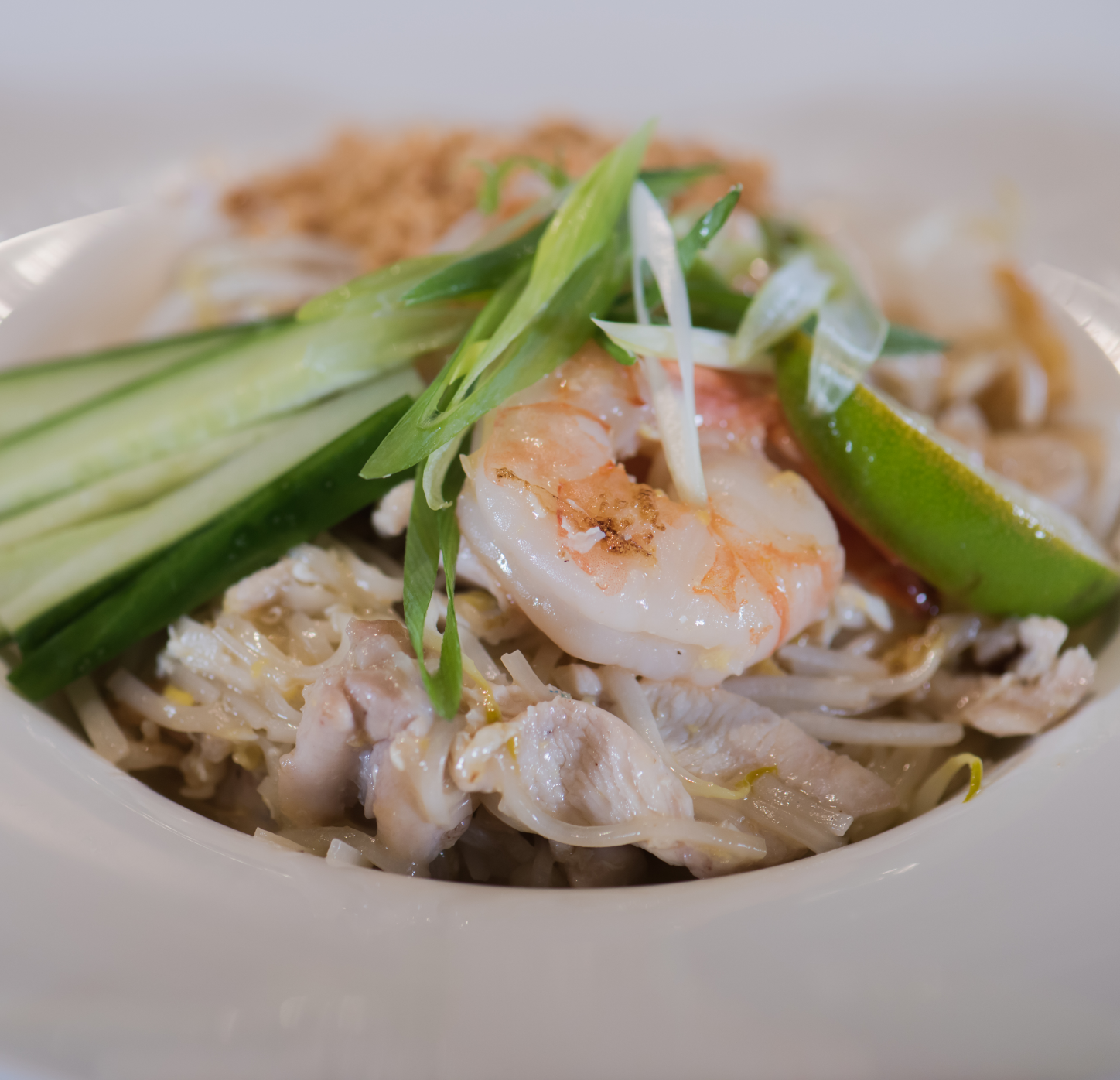 Upclose image of Chef Signaure Pad Thai prepared with house-made authentic sauce, stir-fried rice noodles, egg, preserved radish, tofu, bean sprouts, chicken and shrimp. Garnished with lime, cucumber and peanuts.